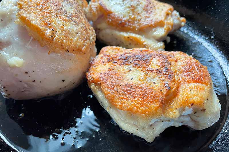 Horizontal image of searing raw poultry pieces in a cast iron skillet.