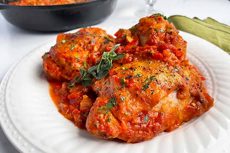 Horizontal image of a white plate with large pieces of cooked poultry covered in tomato sauce with fresh herbs.