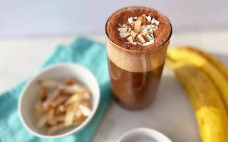 Horizontal image of a cup with a frothy dark chocolate beverage next to a blue towel, a bowl of toasted coconut chips, and a yellow banana