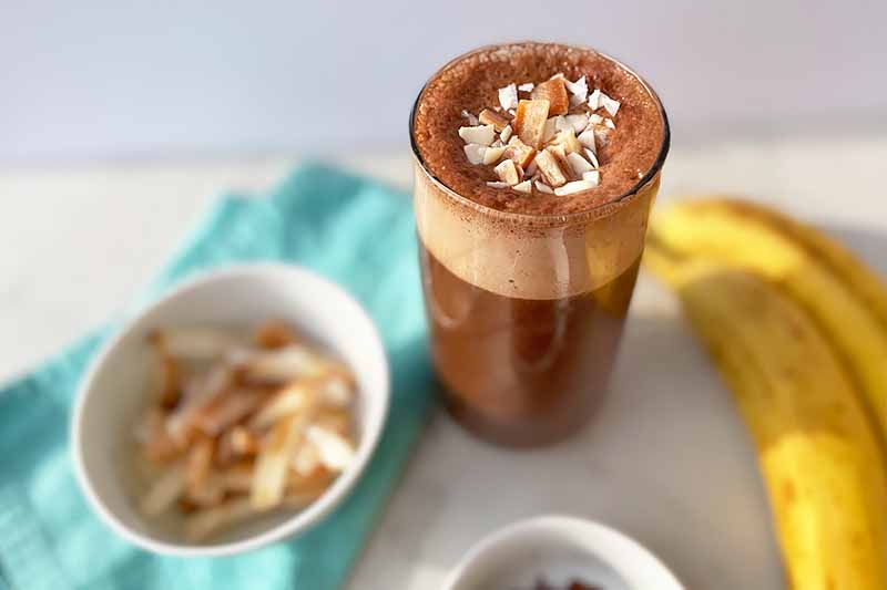 Horizontal image of a cup with a frothy dark chocolate beverage next to a blue towel, a bowl of toasted coconut chips, and a yellow banana