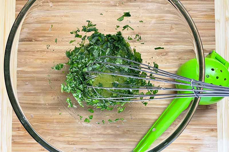 Horizontal image of whisking oil and fresh chopped herbs together in a glass bowl.