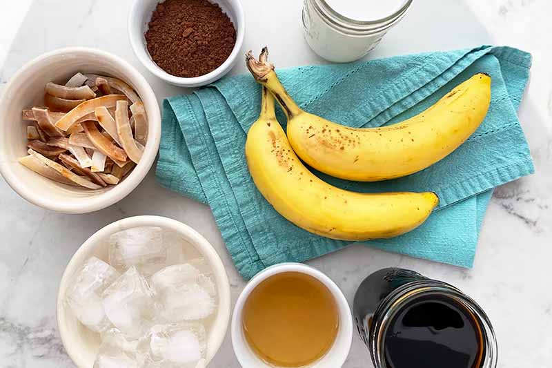 Horizontal image of assorted ingredients in bowls next to two bananas on a blue towel.