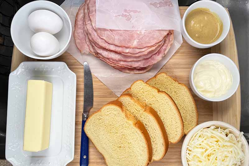 Horizontal image of prepped bread slices, ham slices, butter eggs, cheese, and condiments.