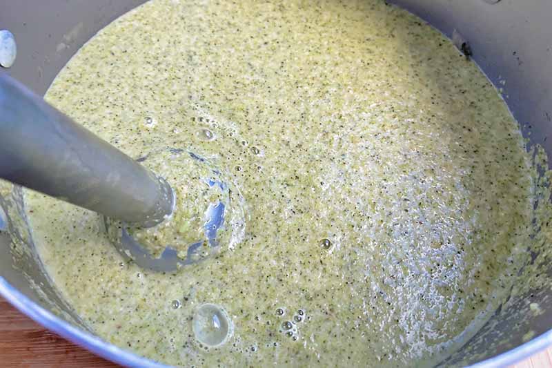 Horizontal image of pureeing a green mixture with an immersion blender in a pot. 