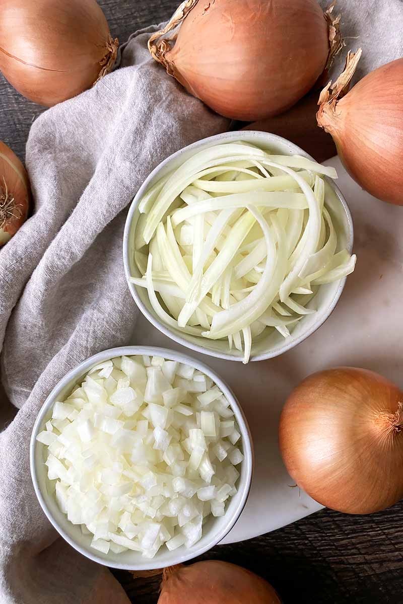 Vertical top-down image of two white bowls filled with sliced and diced white onions.