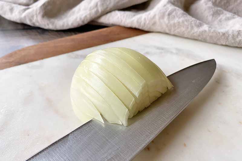 Horizontal image of a knife making a horizontal slice into a white onion with vertical slices in it.