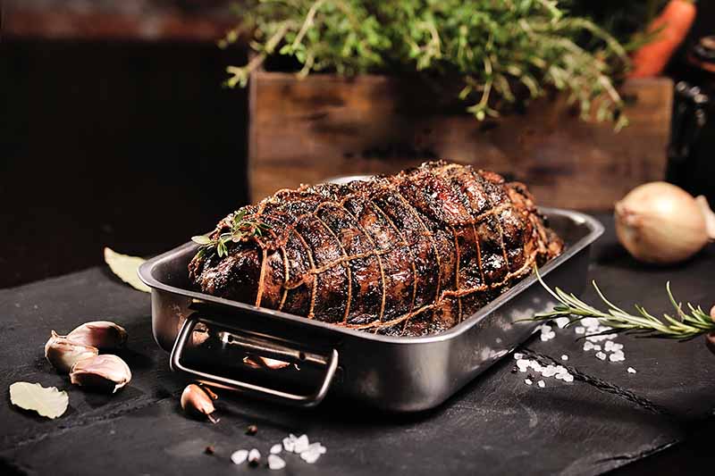 Horizontal image of a large piece of cooked and seasoned meat with kitchen twine in a rectangular skillet surrounded by fresh plants and pieces of garlic cloves.