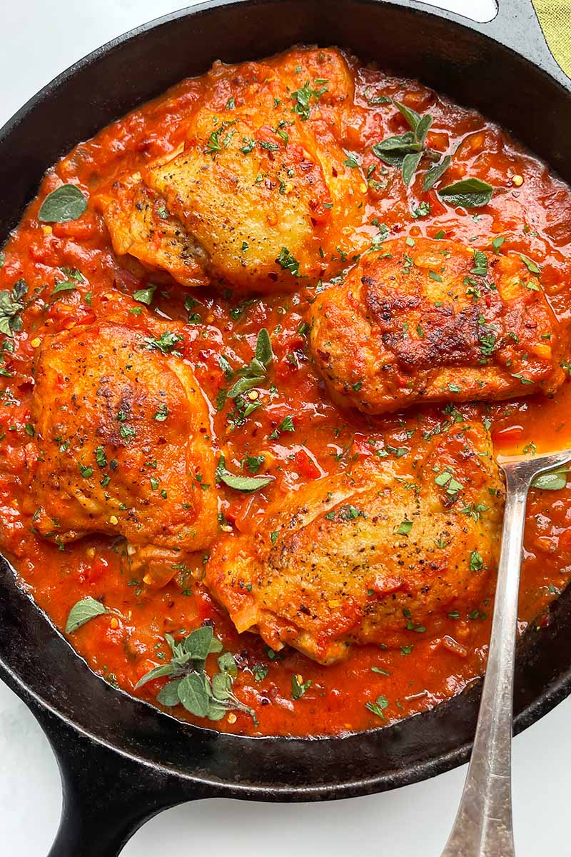Vertical top-down image of four pieces of cooked poultry in a red tomato and herb sauce in a cast iron skillet with a spoon.