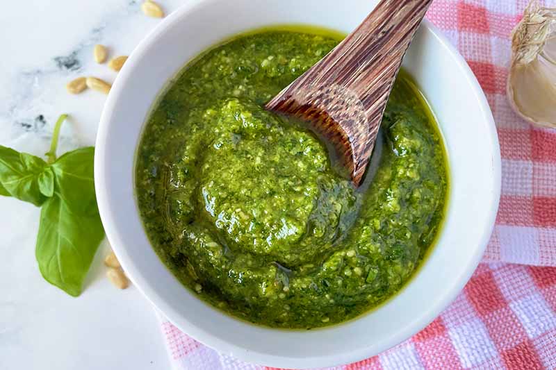 Horizontal image of a spoon holding up some thick green sauce over a white bowl on a red checkered towel.
