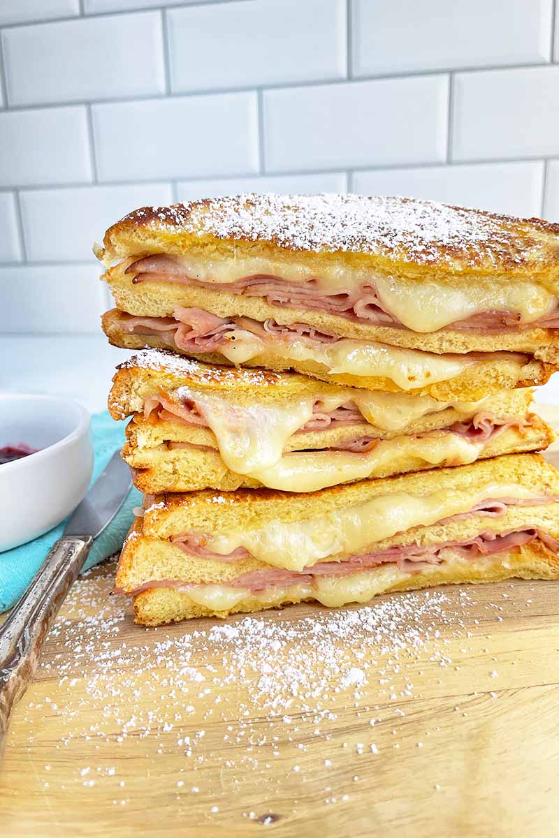 Vertical image of stacks of sandwiches with ham slices and melted cheese on a wooden cutting board.