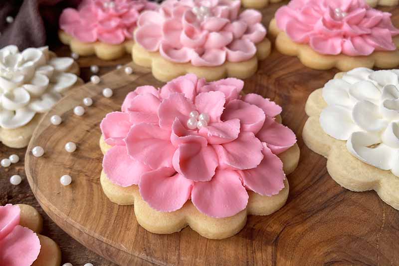 Horizontal close-up image of a pink floral cookie on a wooden surface next to white sprinkles.