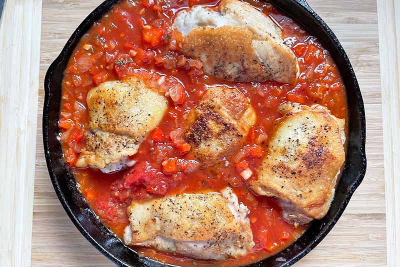 Horizontal image of simmering pieces of poultry in a sauce in a cast iron skillet.