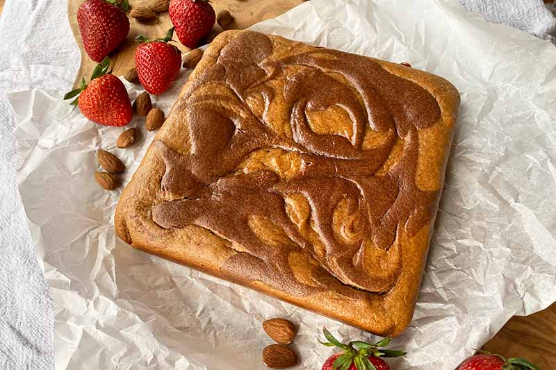 Horizontal image of a square cake with a brown swirl on top on a parchment paper next to strawberries.