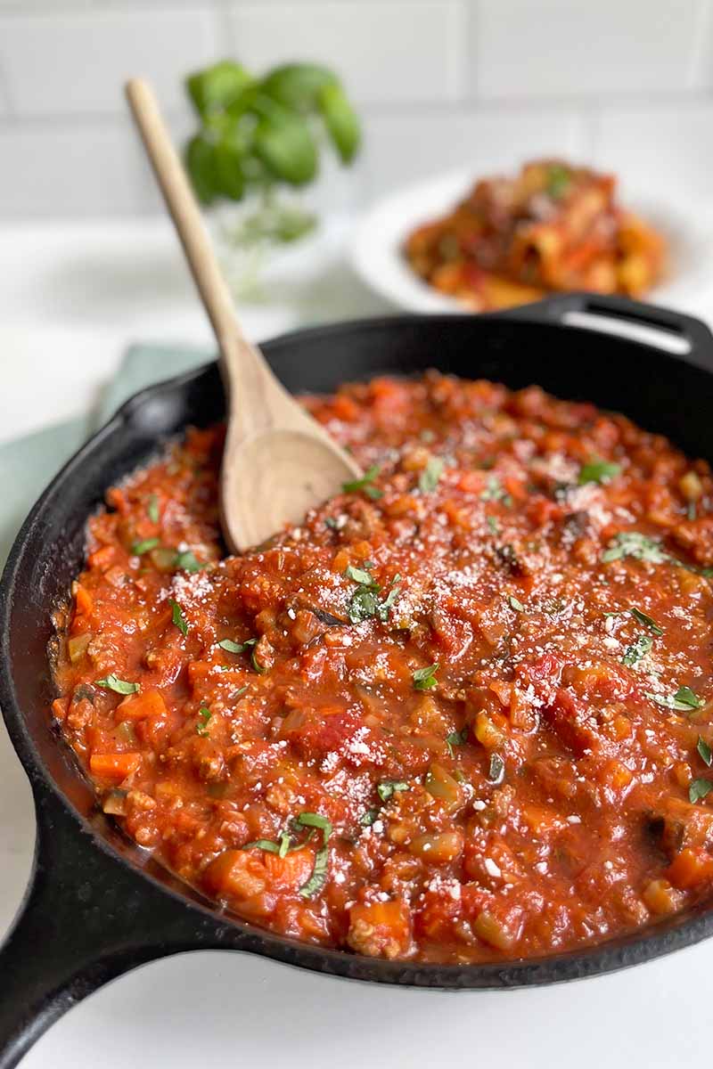Vertical image of a thick tomato sauce finished with chopped basil in a cast iron skillet stirred with a wooden spoon.