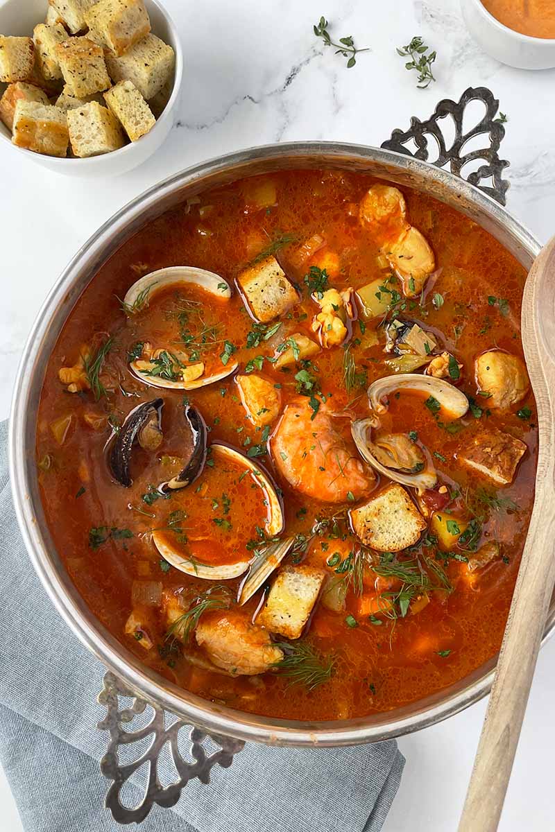 Vertical image of a large pot full of tomato broth with chunks of vegetables and whole shellfish next to a white bowl with bread cubes.