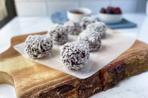 Horizontal image of two rows of small chocolate mounds on parchment on a wooden cutting board in front of bowls of ingredients on a blue towel.