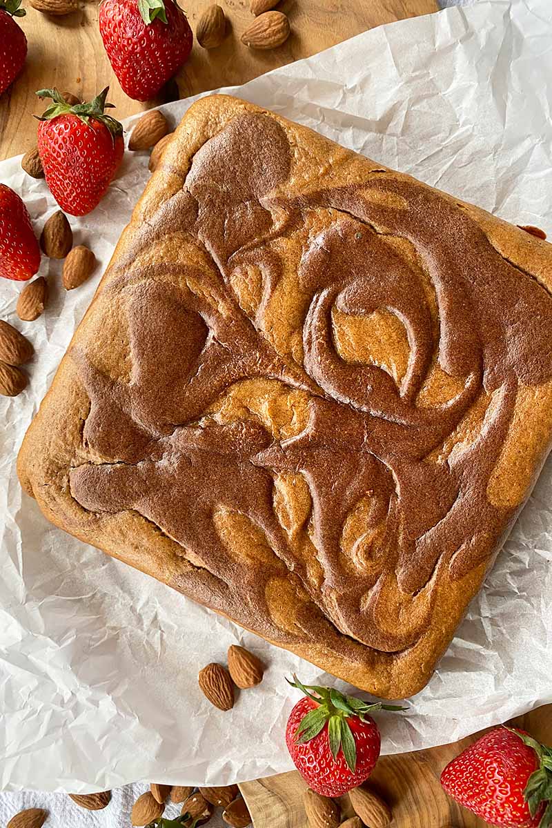 Vertical image of a square baked good with dark brown swirls, on white parchment paper surrounded by strawberries and nuts.