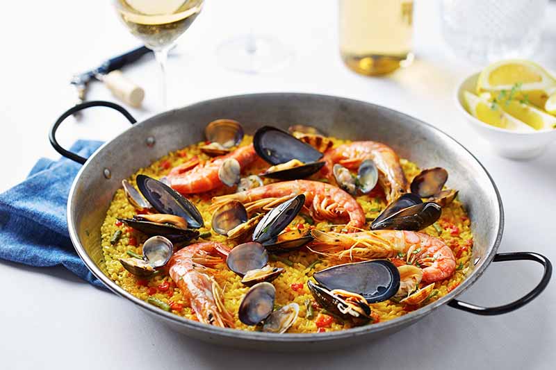 Horizontal image of paella in a pan on a blue napkin next to glasses of wine.