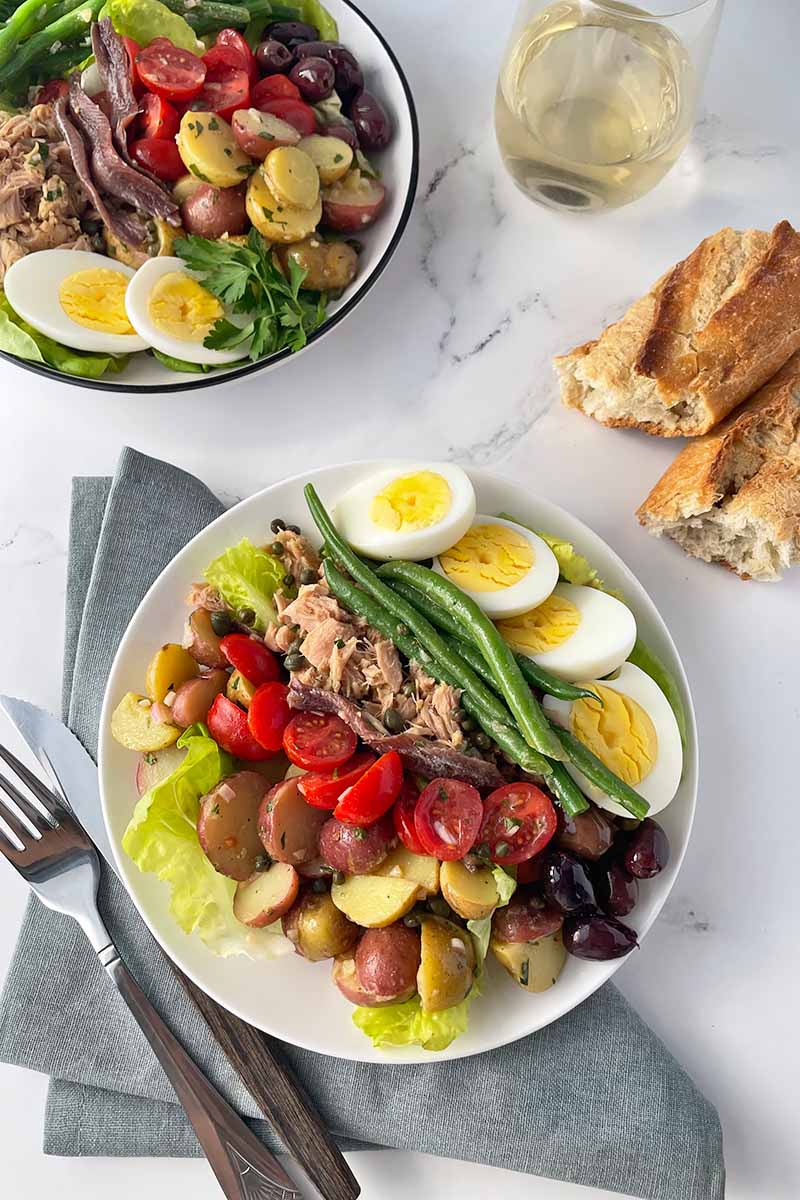 Vertical image of two plates with assorted vegetables, seafood, and slices of hard-boiled eggs on gray napkins next to silverware, a glass of white wine, and pieces of bread.