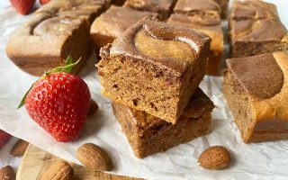 Horizontal image of dessert squares with a brown swirl on top on parchment paper next to strawberries and nuts.