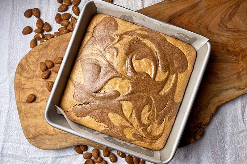 Horizontal image of a baked good with a cinnamon swirl on the surface in a metal pan next to almonds.