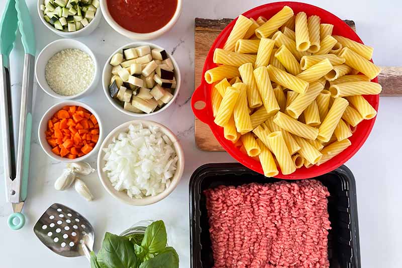 Horizontal image of assorted ingredients in bowls, pasta in a red colander, and raw beef in a black container.