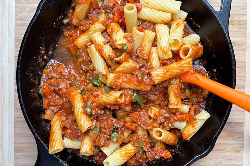 Horizontal image of tossing cooked rigatoni with sauce in a cast iron skillet.