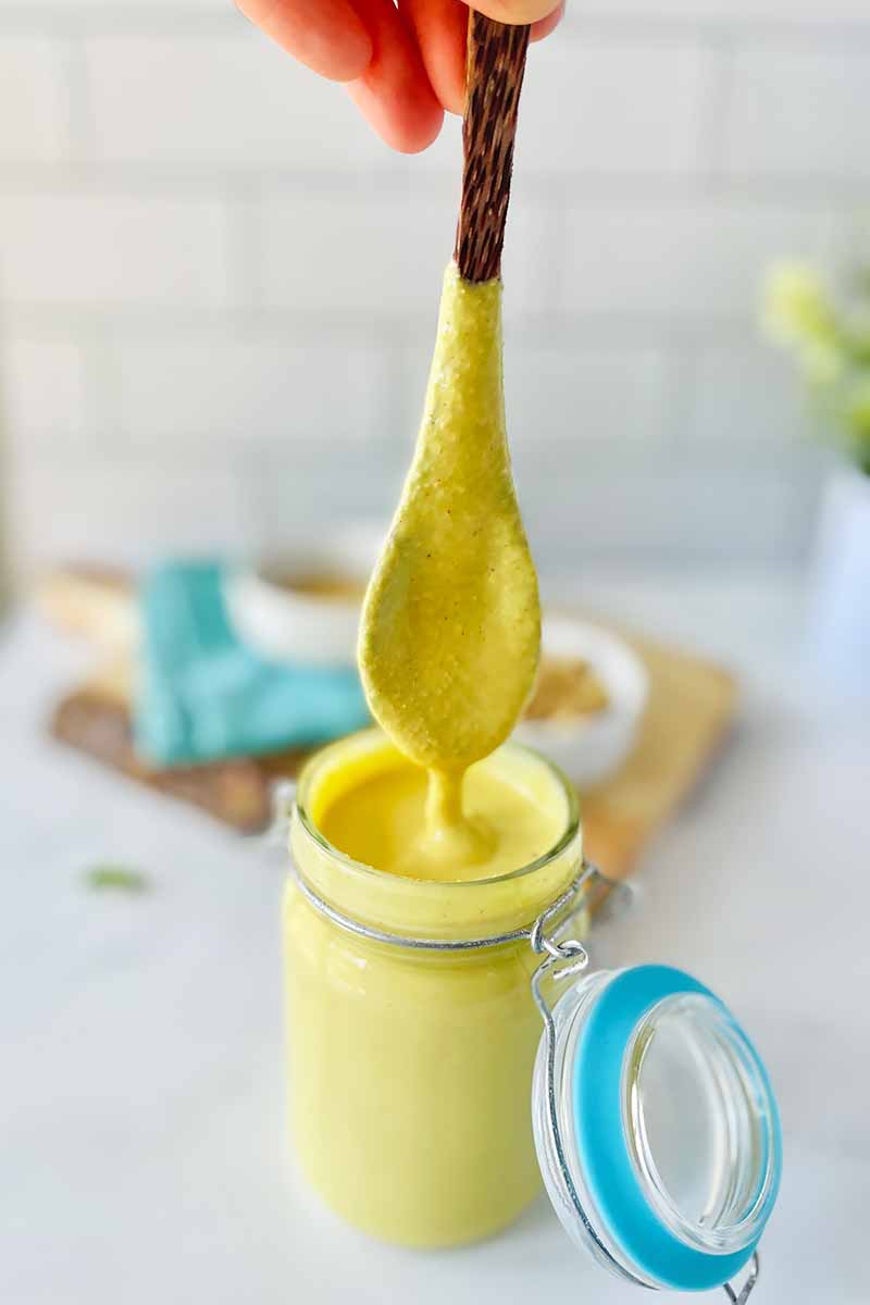 Vertical image of a hand holding a spoon dipped in a thick yellow mixture in a jar in front of a wooden cutting board with a bowl on a blue towel.