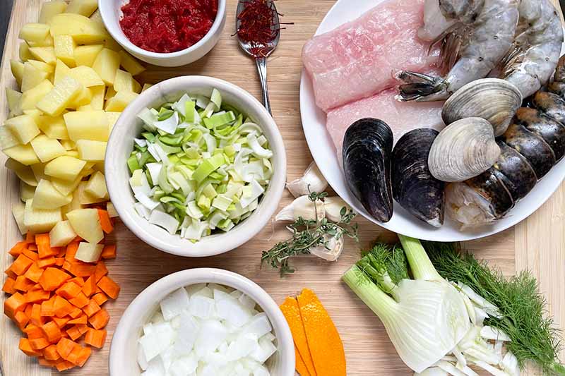 Horizontal image of assorted prepped vegetables, fish and shellfish, and aromatics, with some in white bowls.