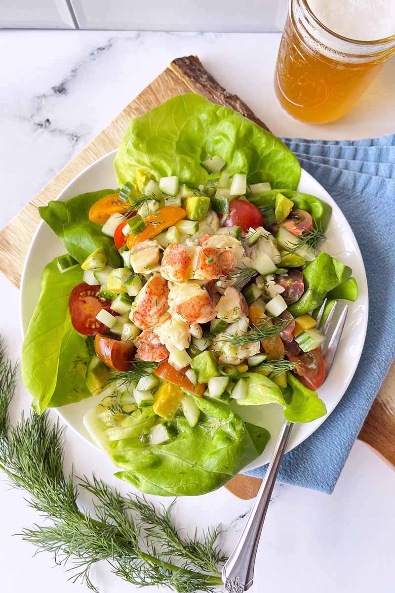 Vertical top-down image of bib lettuce with a seafood and fresh vegetable mixed salad on top, with fresh herbs, a blue napkin, and a glass of beer next to it.