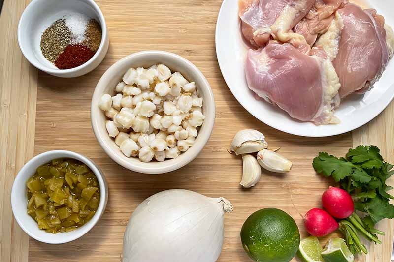 Horizontal image of assorted ingredients and raw poultry thighs in white bowls on a wooden cutting board next to fresh produce.