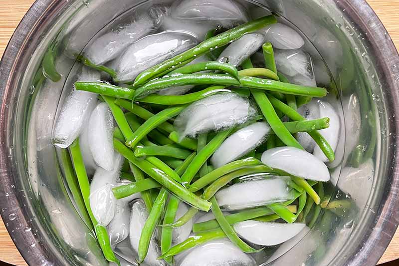 Horizontal image of a metal bowl with green beans and ice water.