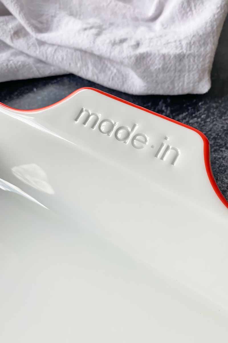 Vertical image of a handle of a casserole with a red rim next to a white towel.