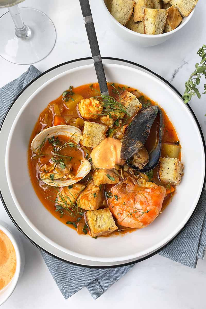 Vertical image of a white bowl filled with a shellfish soup in a tomato broth on a gray napkin with a spoon.