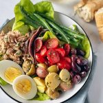 Horizontal image of assorted fresh vegetables, olives, canned fish, and slices of hard-boiled eggs on a white plate next to chunks of bread.