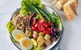 Horizontal image of assorted fresh vegetables, olives, canned fish, and slices of hard-boiled eggs on a white plate next to chunks of bread.