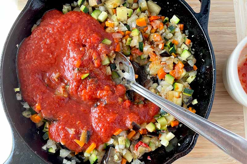 Horizontal image of adding tomato sauce to a cast iron skillet with cooked assorted vegetables.