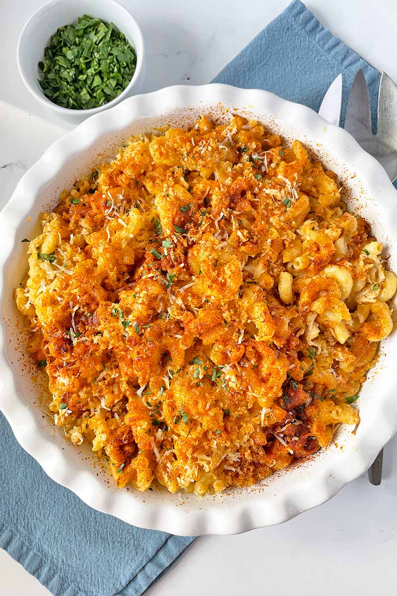 Vertical top-down image of a pasta casserole in a white pie dish topped with buttered toasted breadcrumbs on a blue napkin next to chopped herbs.