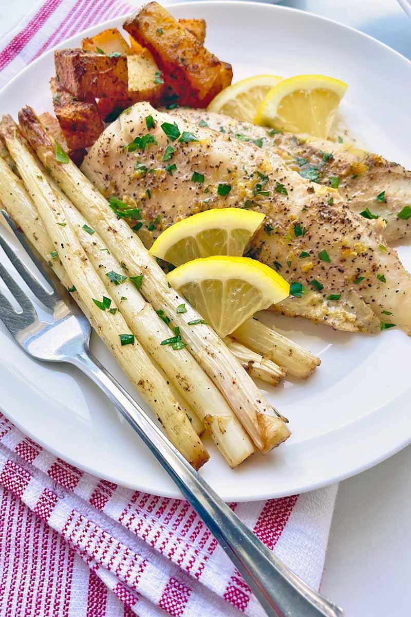Vertical close-up image of white asparagus, roasted potatoes, fish fillets, and lemon slices on a white plate next to silverware and a red napkin.