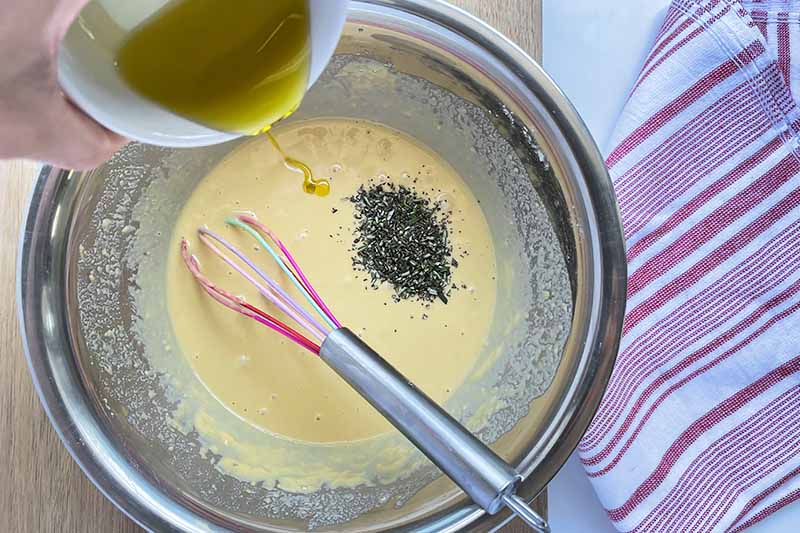 Horizontal image of pouring oil into a yellow batter with a small mound of chopped herbs.