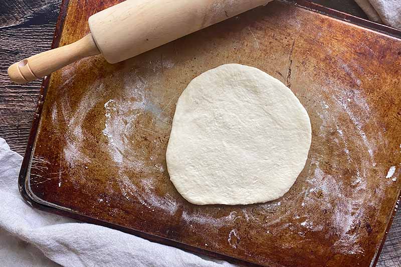 Horizontal image of a flattened disc of dough on a lightly floured pan next to a rolling pin.