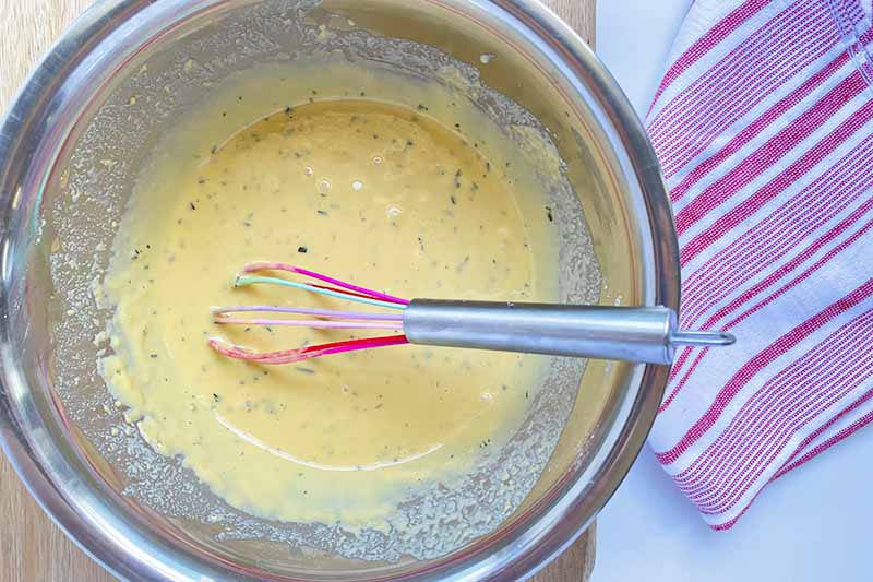 Horizontal image of a thick yellow batter with crushed rosemary in a metal bowl. with a whisk.