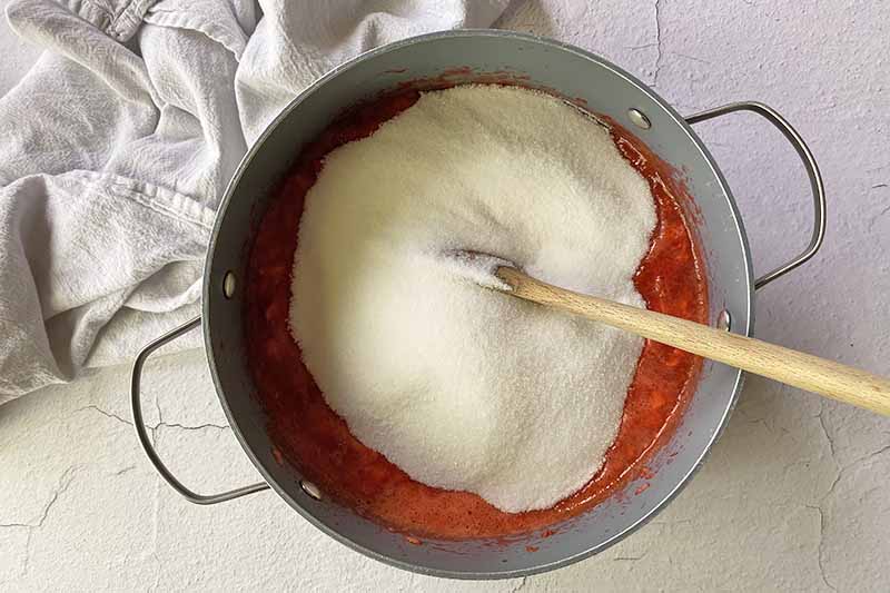 Horizontal image of a large pile of granulated sugar over a red liquid in a pot stirred with a wooden spoon.
