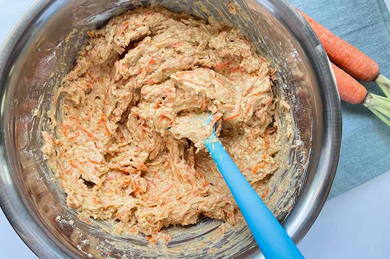 Horizontal image of mixing together a batter with shredded orange vegetables in a metal bowl.