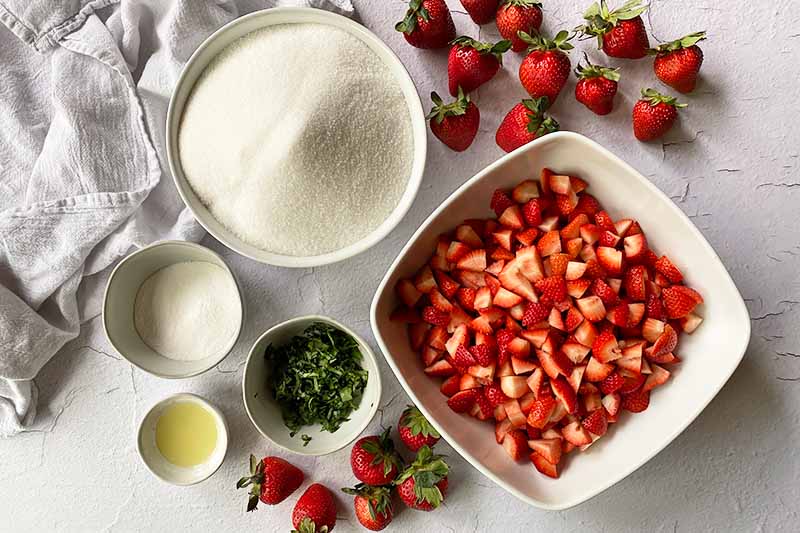 Horizontal image of bowls of sugar, strawberries, chopped aromatics, and other ingredients.