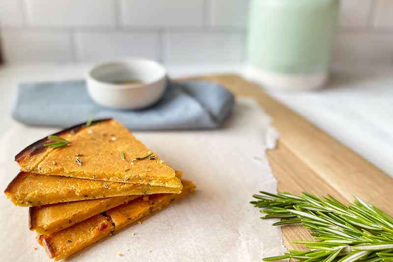 Horizontal image of triangular pieces of flatbread on a wooden cutting board with parchment paper, next to fresh herbs, a white bowl, and a blue towel.