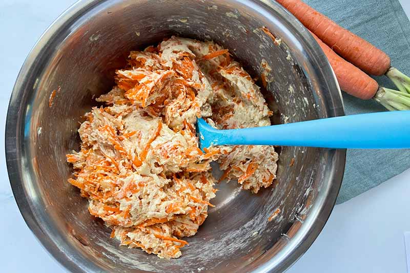 Horizontal image of folding together thick wet ingredients and shredded orange vegetables in a metal bowl.