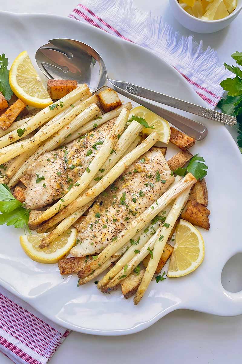 Vertical image of a platter full of white asparagus, fish fillets, and lemon slices next to a metal spoon.
