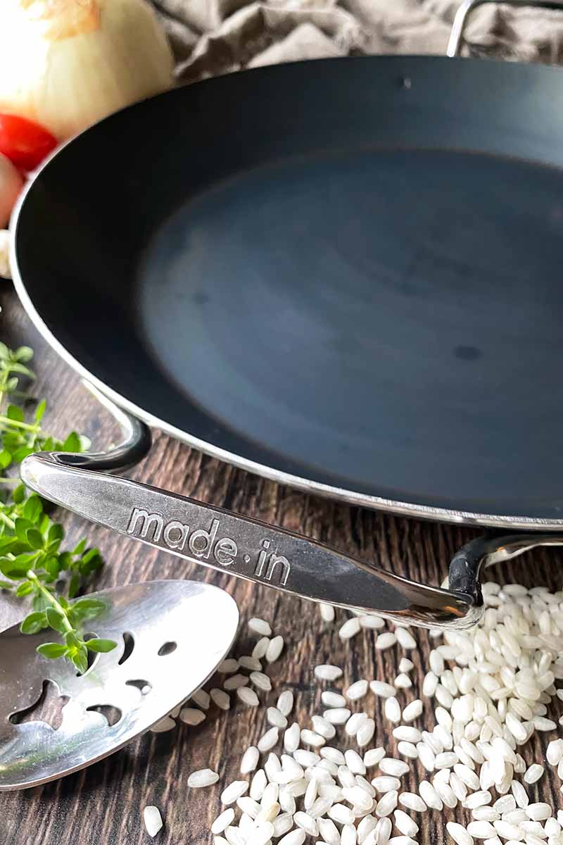 Vertical image of an empty shallow cookware with silver handles next to rice, a metal spoon, and produce.