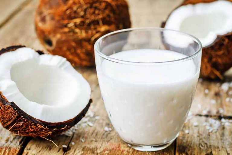 Horizontal image of a glass of white liquid next to halved coconut on a wooden surface.
